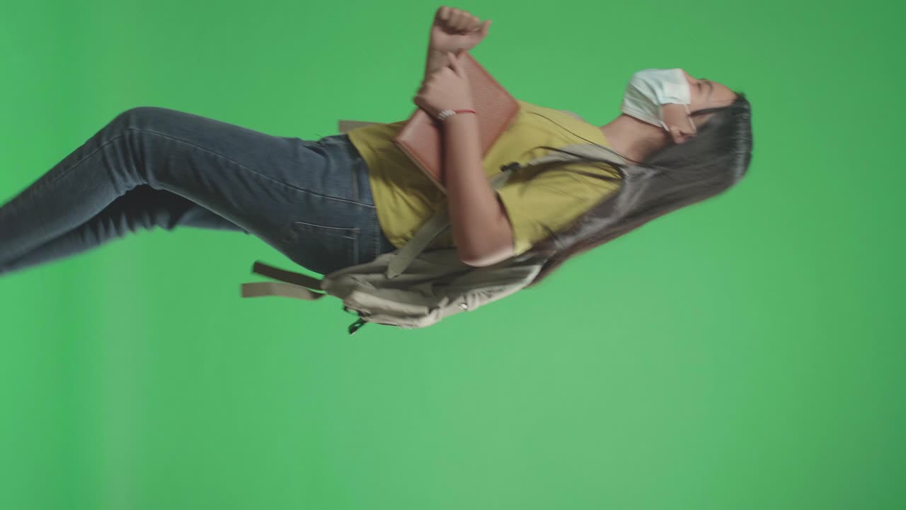 vista lateral de una estudiante asiática con una máscara, sosteniendo un libro mientras corre a la escuela en una pantalla verde