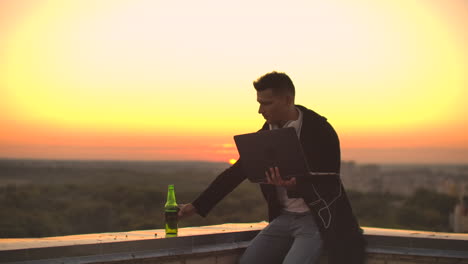 A-young-man-sitting-on-the-edge-of-the-roof-with-a-laptop-and-a-beer-working-typing-on-a-laptop