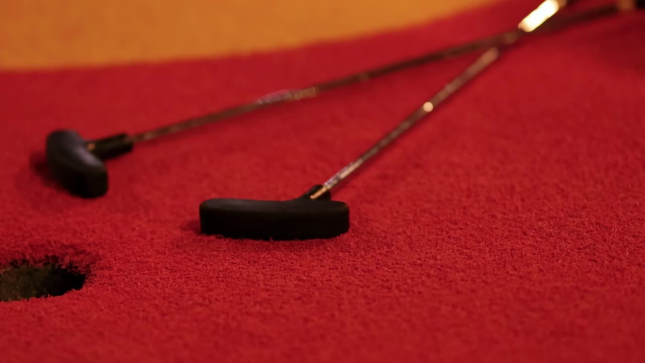 A red golf ball narrowly misses the hole on a vibrant red mini golf course with putters nearby.