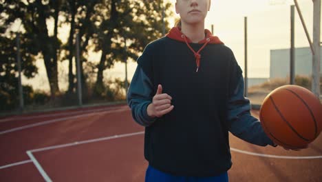 A-close-up-portrait-of-a-blonde-girl-in-a-sports-uniform-who-throws-an-orange-ball-from-hand-to-hand-on-a-red-basketball-summer-street-court-in-the-morning