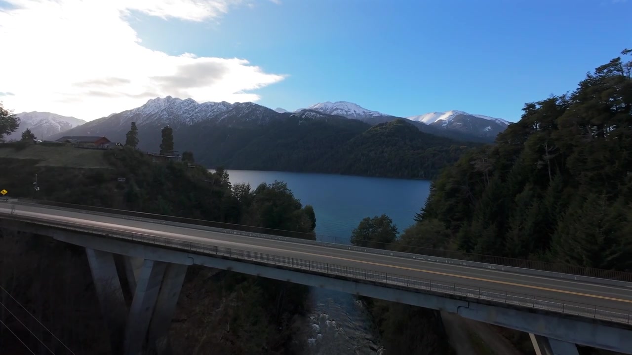 Vista del ponte autostradale sul Rio Correntoso e cime innevate e lago a Villa La Angostura, Neuquén. Argentina. Passaggio aereo ascendente in avanti in una mattina d'inverno parzialmente nuvolosa