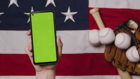 Person-Holding-Green-Screen-Mobile-Phone-Above-Baseball-Still-Life-With-Bat-Ball-And-Catchers-Mitt-On-American-Flag