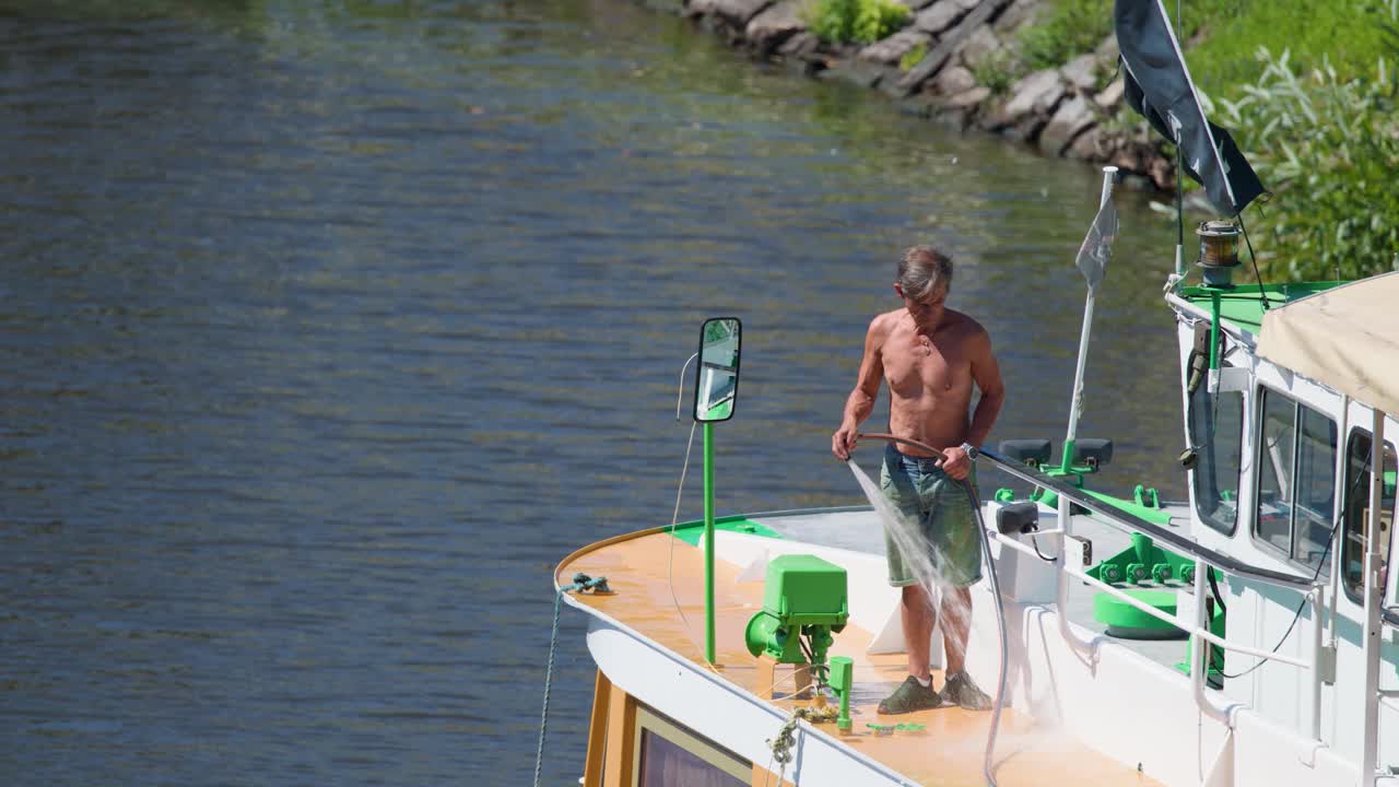 Shirtless man washes boat deck with hose at marina, bright daylight, steady camera, relaxed mood