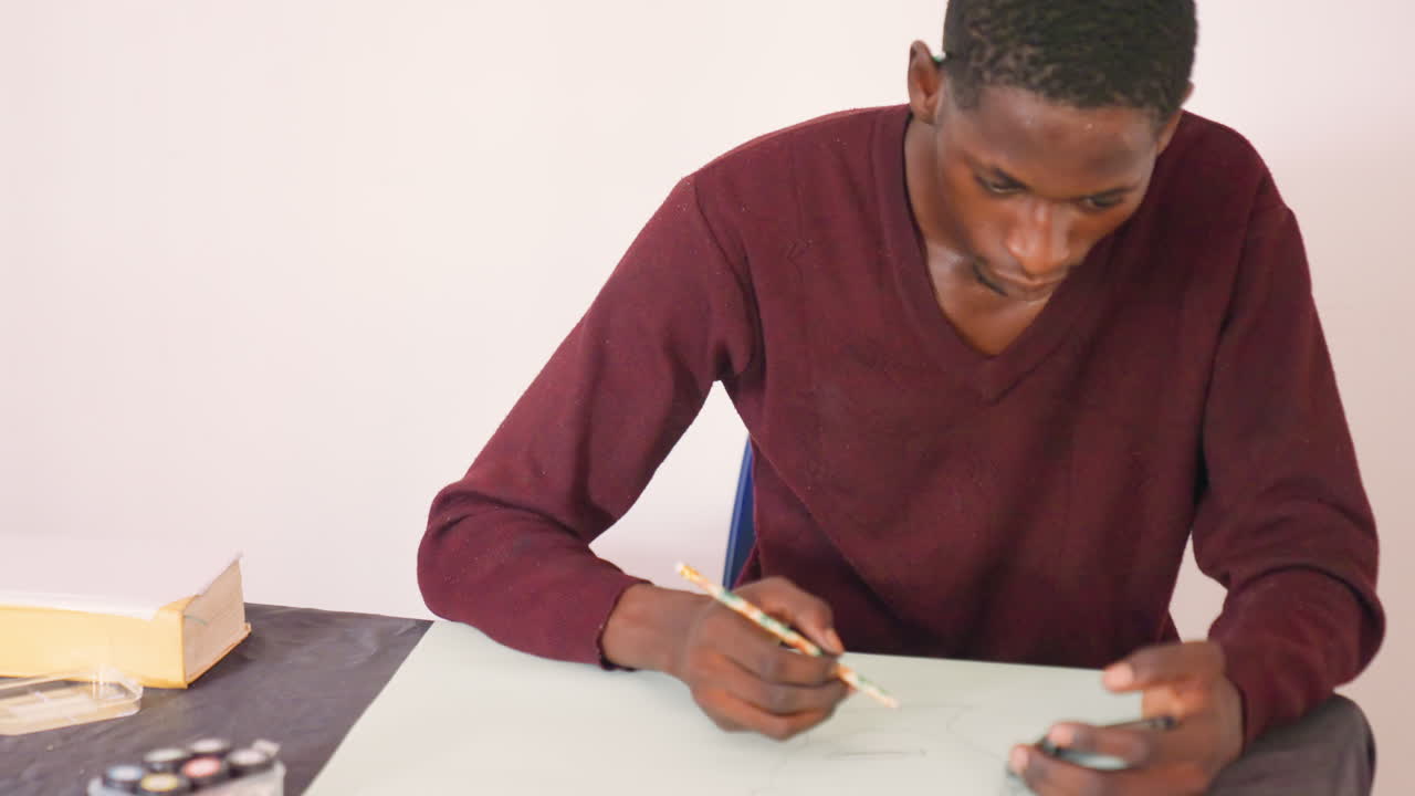Artista uomo nero che dipinge su carta, uno studente concentrato che disegna con pennello e matita, indossa un maglione bordeaux, seduto a un tavolo bianco con tavolozza e strumenti nelle vicinanze. Mani in primo piano, sotto una luce calma da studio, un creativo concentrato.