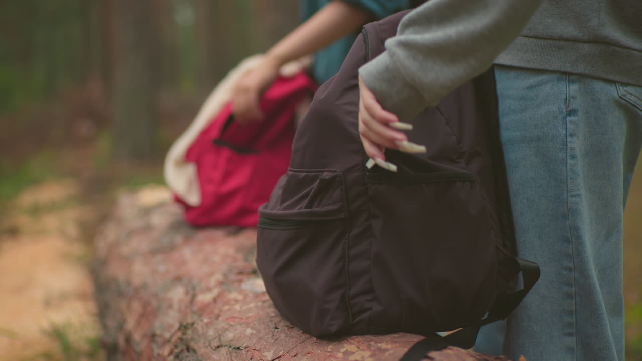 Close-up side view of two backpacks resting on a fallen tree in a tranquil forest setting, they both open their bag preparing for rest, with background featuring greenery
