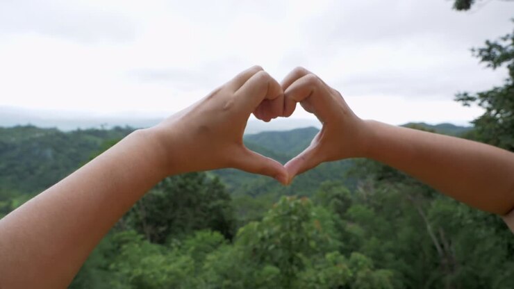 Two hands joined together to form a heart shape, a symbol of love and affection against a natural background of mountains and trees