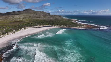 Hermosa-Toma-Aérea-Escénica-De-Una-Playa-Con-Colinas-Rocosas,-Aguas-Verdes-Marinas,-Olas-Perezosas-Y-Cielo-Parcialmente-Nublado-Cerca-De-Ciudad-Del-Cabo-Sudáfrica