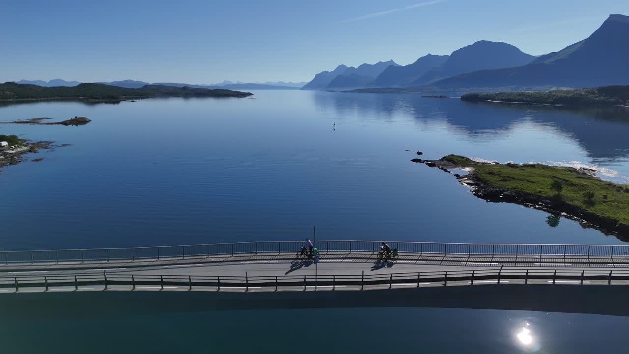Aerial view of bikers standing on a scenic bridge in Norway, admiring tranquil fjord reflections and dramatic mountain landscape at sunrise