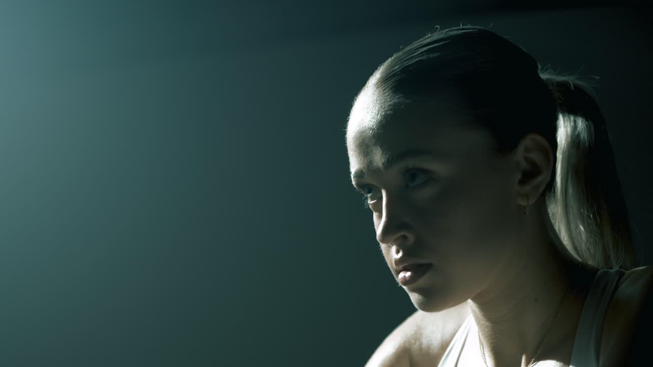 Side view of a woman cycling in a dark studio, backlit with soft cinematic light as the camera moves to the front, revealing her focused eyes and deep breathing during intense training effort