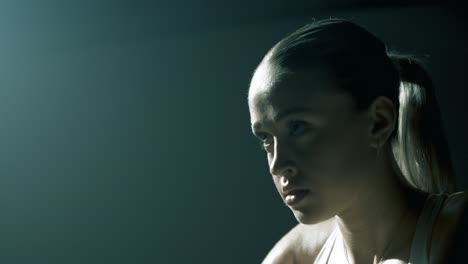 side view of a woman cycling in a dark studio, backlit with soft cinematic light as the camera moves to the front, revealing her focused eyes and deep breathing during intense training effort