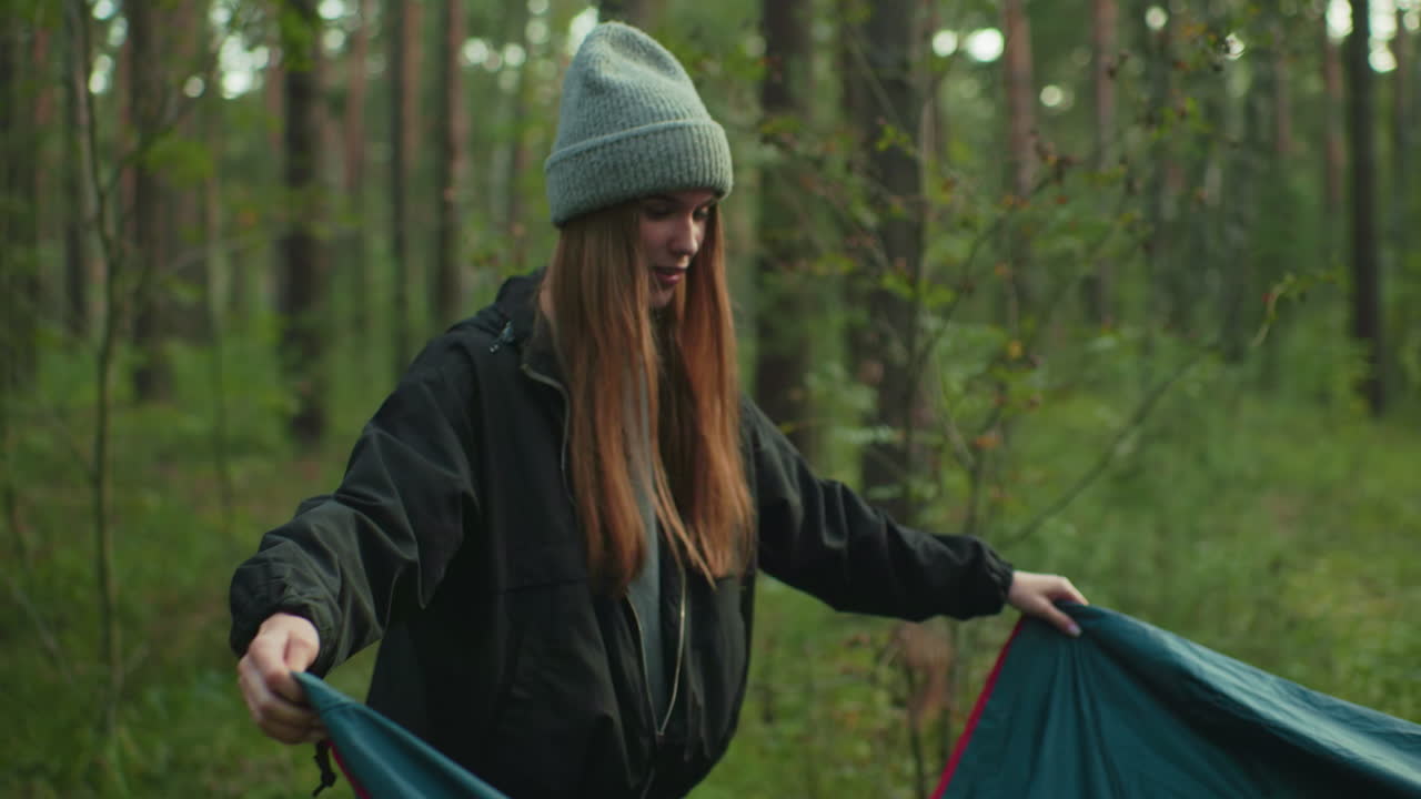 Young woman lifts tent cover in forest campground with partner helping behind pitched tent, smiling and preparing gear on grassy ground beneath tall trees during serene outdoor adventure