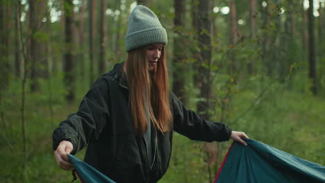 young woman lifts tent cover in forest campground with partner helping behind pitched tent, smiling and preparing gear on grassy ground beneath tall trees during serene outdoor adventure