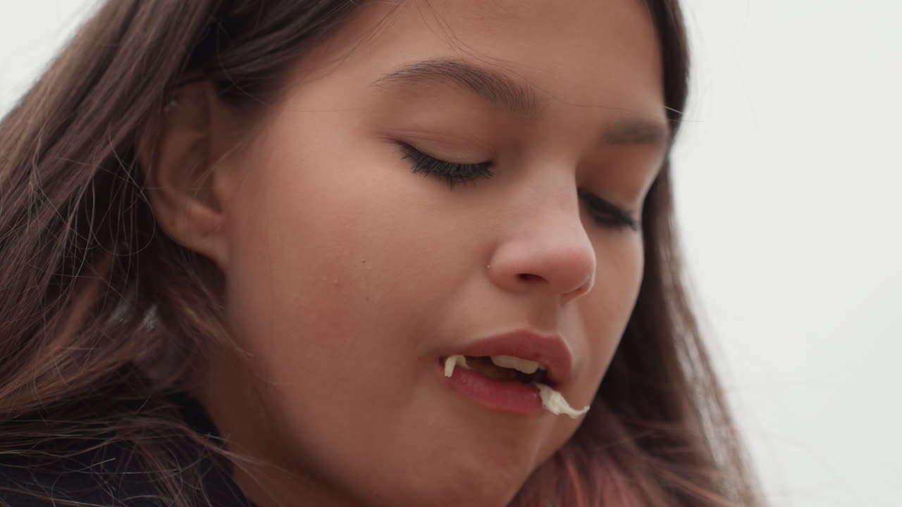teen latina student pensive windblown portrait, downcast gaze and lips pressed, hair across face, overcast sky, subtle chewing at sandwich, quiet urban backdrop, reflective mood and solitary waiting