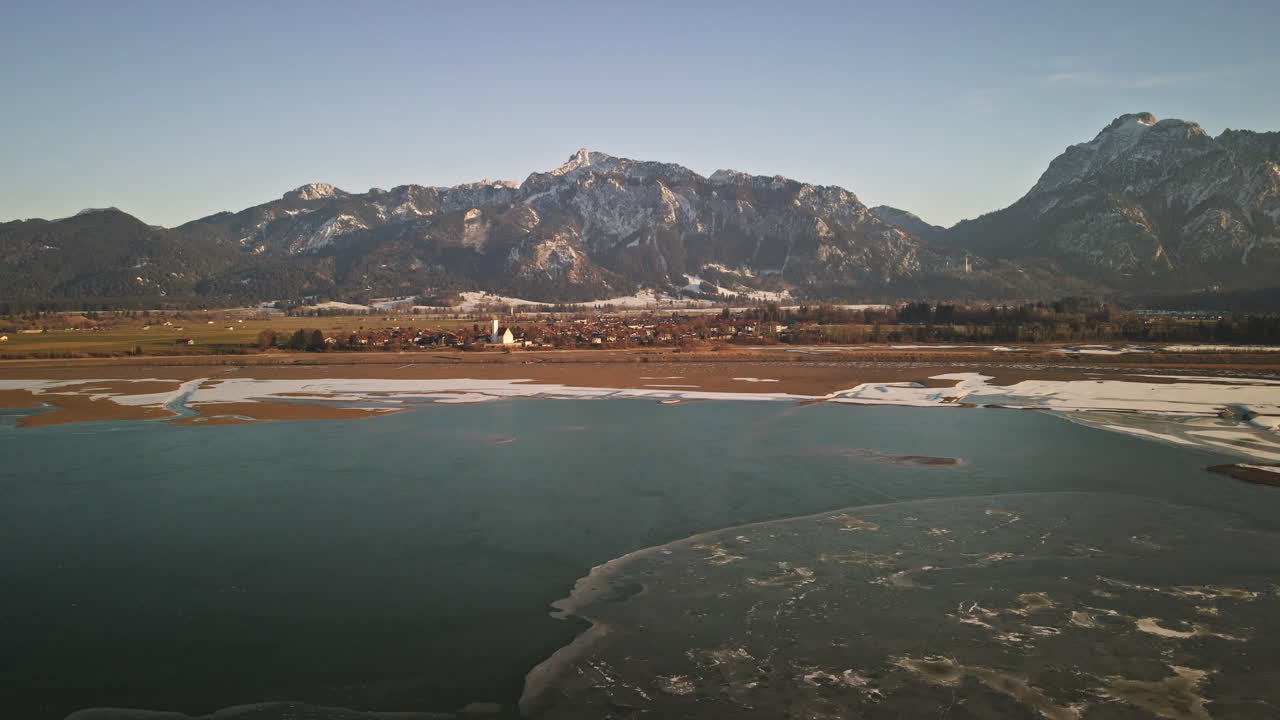 Camera glides forward along a narrow sandbar where snow and ice meet deep blue water, with Bavarian mountains and a winter village in the distance