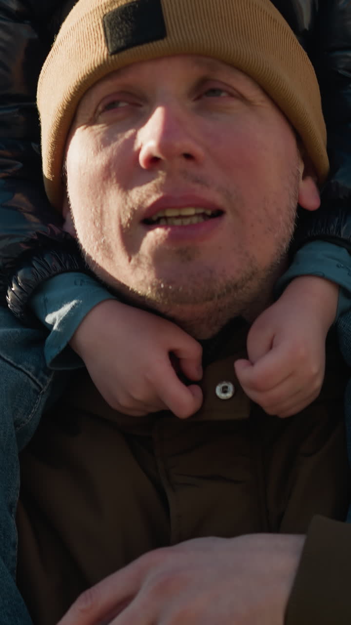 un primer plano de un padre llevando a su hijo en sus hombros, con el padre sosteniendo la pierna de su hijo con seguridad, el padre está hablando con su hijo, con una vista borrosa de los árboles en el fondo