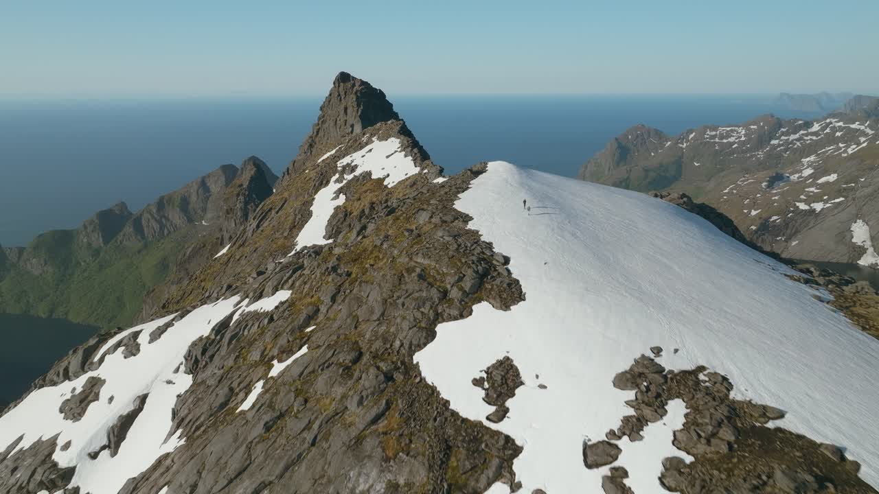 Luftdroneflyvning over Munken-toppen i Lofoten, Norge, hvor to vandrere krydser en sneplet på et barskt granitbjerg under en klar himmel.