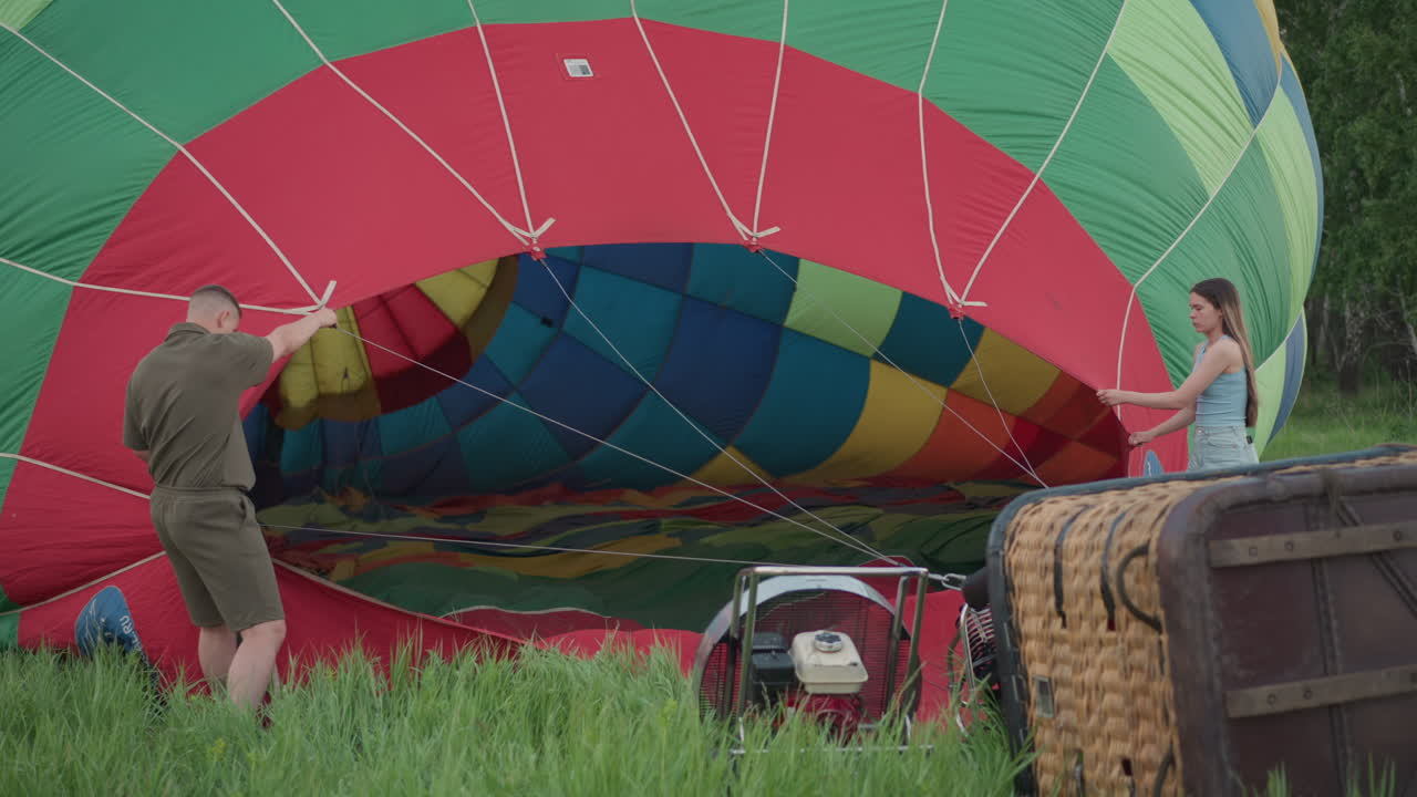 man and woman holding hot air balloon envelope in vast green field at sunset preparing for launch with wind inflating vibrant fabric while crew readies burner and basket under pastel sky
