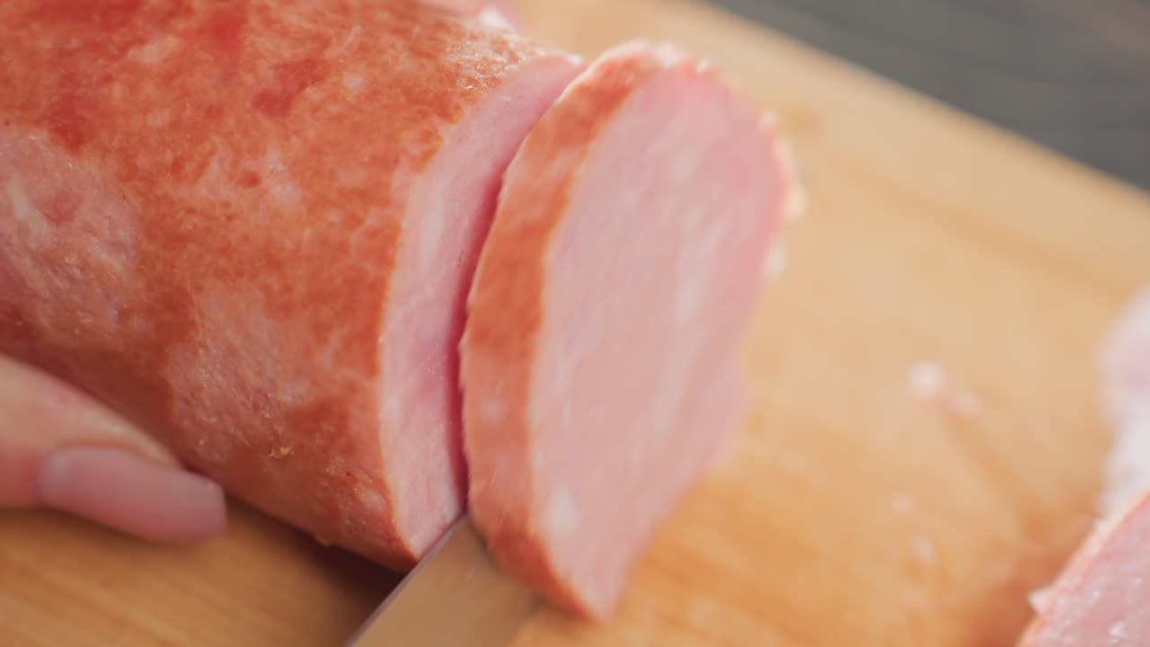 close up hands slicing pink pork sausage on wooden cutting board using sharp kitchen knife under natural light, showing clean cut surface and smooth meat texture during food preparation process