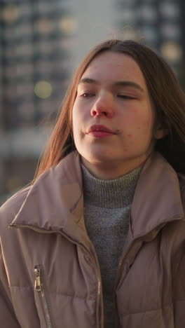 a young woman wearing a peach jacket is seen sitting in an urban park with a contemplative expression on her face. the blurred background, featuring tall buildings and evening lights