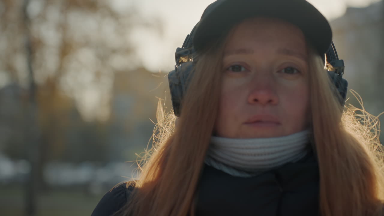 Donna caucasica controluce in un parco, capelli rossi con lentiggini, cuffie e berretto, sciarpa avvolta, fiato visibile nella luce fredda, sguardo contemplativo verso la strada, ritratto cinematografico all'ora d'oro, d'atmosfera