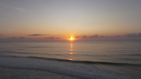 aerial backwards shot showing sunset at horizon behind ocean with calm waves at gillards' beach australia