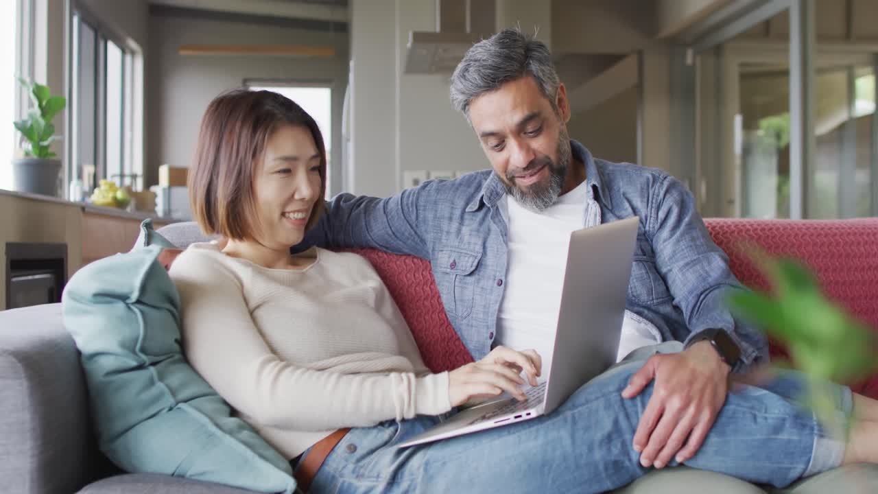 una pareja feliz y diversa sentada en la sala de estar usando una computadora portátil y hablando