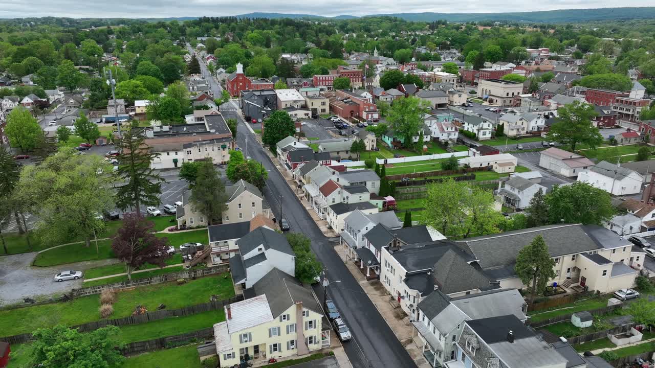 Main street of small american town during cloudy day in Pennsylvania. Green trees and green yards of homes along road. Aerial wide shot. Historic city with church and buildings. Manheim, USA.
