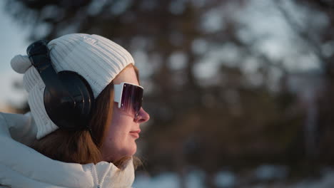 snow ground with silhouetted trees and sun peeking through branches, city buildings blurred, back lit winter scene, soft glow over serene urban nature, young woman grooving to music