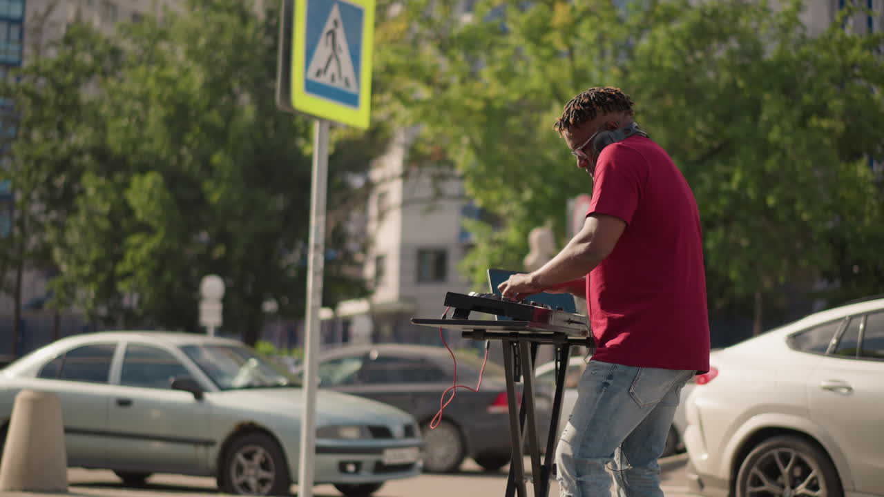Closeup DJ adjusting headphones and mixer, concentrated expression as he finetunes levels on portable controller with blurred city cars and crosswalk sign behind red shirt and casual jeans, warm