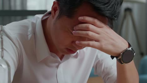 close up of drunk asian man with vodka glass holding the temples while sitting on the chair at home