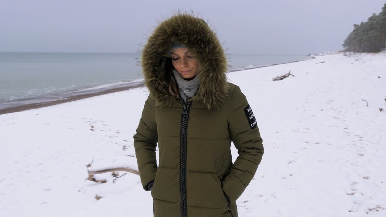 녹색 겨울 옷을 입은 젊은 여성이 발트해 해변의 모래사장을 따라 걷고, 눈 덮인 겨울날, 핸드헬드 추적 샷