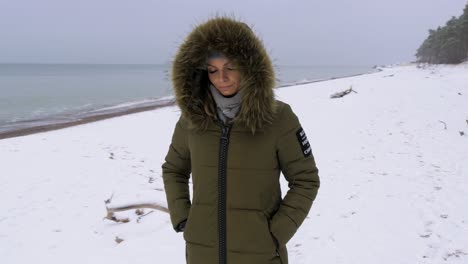 Young-woman-in-green-winter-clothes-walks-along-the-sandy-shore-of-the-Baltic-sea-beach,-overcast-snowy-winter-day,-handheld-tracking-shot