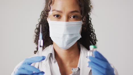 vídeo del retrato de una mujer médica biracial con máscara facial sosteniendo un vial de vacuna sobre un fondo blanco