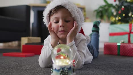 surprised caucasian boy wearing santa hat lying on floor, watching snow globe