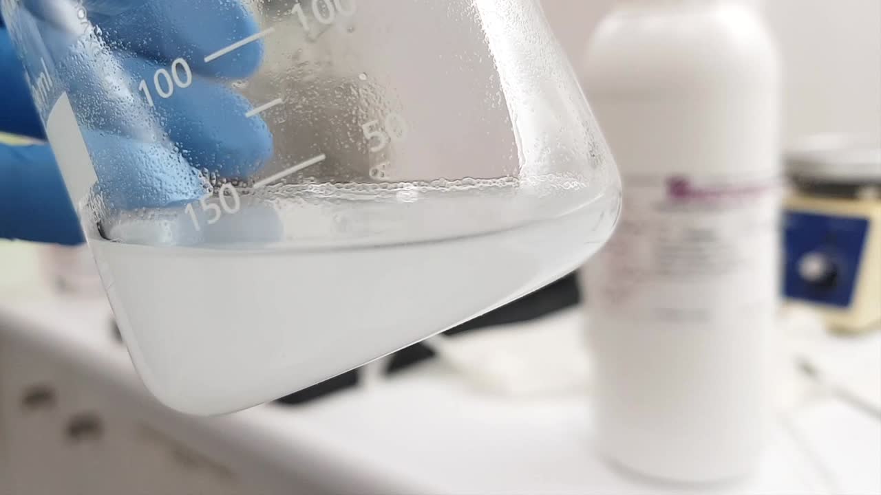 A close-up shot of a laboratory beaker filled with a turbid liquid. A gloved hand holds the beaker carefully, highlighting the experiment taking place in a professional chemistry lab.