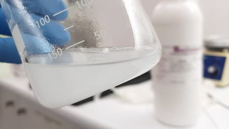 a close-up shot of a laboratory beaker filled with a turbid liquid. a gloved hand holds the beaker carefully, highlighting the experiment taking place in a professional chemistry lab.