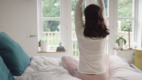 Vista-Trasera-De-Una-Mujer-Asiática-Madura-En-Pijama-Sentada-En-La-Cama-Meditando-En-Pose-De-Yoga---Filmada-En-Cámara-Lenta