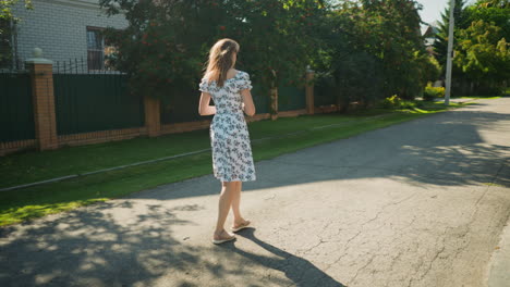 calm girl walking gracefully in floral dress on sunny day along residential street lined with brick fences, greenery, and trees casting gentle shadows on peaceful paved road