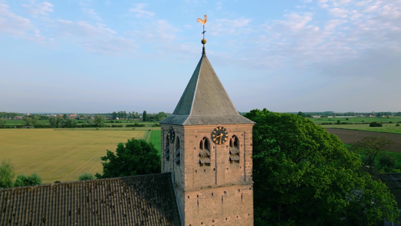 Vista aerea ravvicinata della storica torre della Chiesa Vecchia di Oosterbeek nei Paesi Bassi, illuminata dalla luce dell'ora d'oro. Presenta intricati dettagli architettonici, quadranti dell'orologio e una prominente banderuola segnavento.