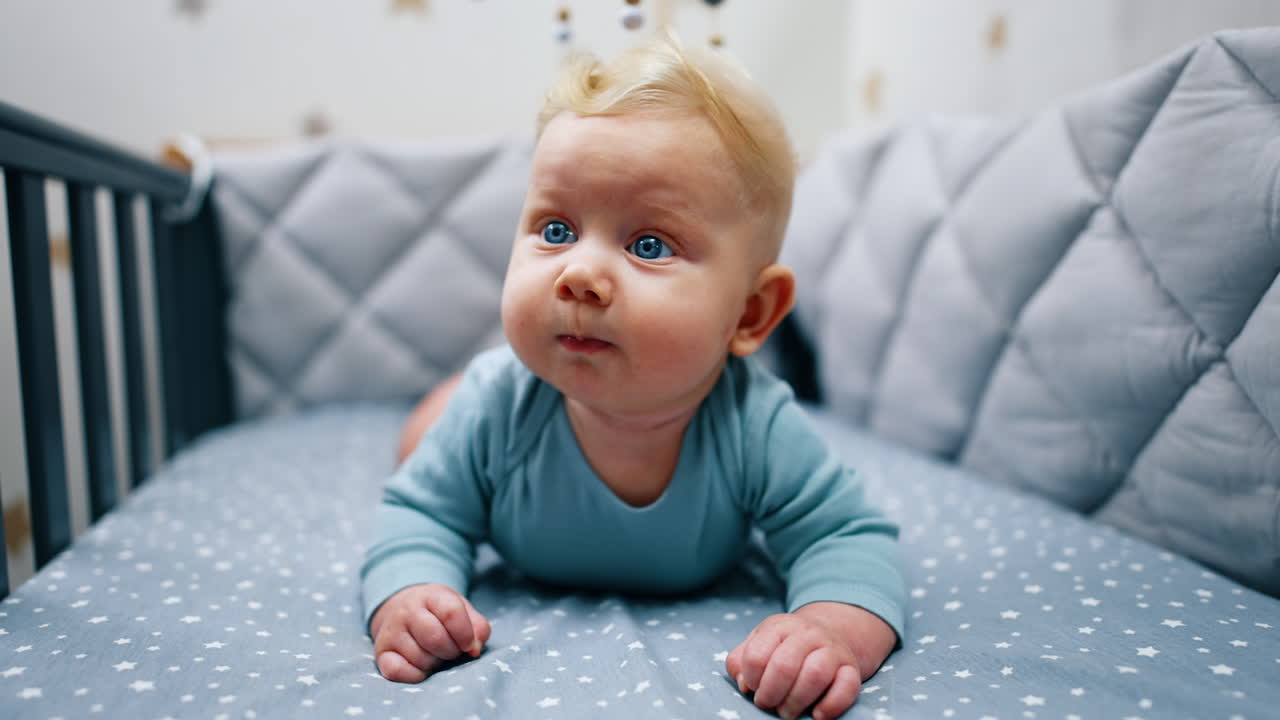 Adorable blue-eyed blond baby lies in the crib. Cute calm infant boy looking surprised around. Close up.