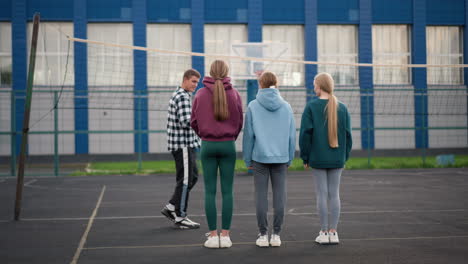 rear view of young volleyball players standing in sportswear, attentively listening to coach instructing them on an outdoor court, blue building and net in background