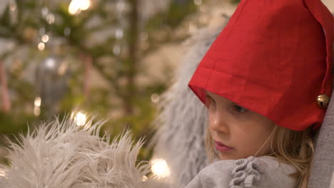 linda niñita con gorro de papá noel sentada frente a un árbol de navidad, de cerca