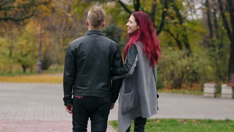 Bacl-view-of-romantic-young-couple-walking-in-autumn-park-during-the-day.-Back-view-of-young-blonde-man-in-leather-jacket-and-his-girlfriend-talking-and-spending-time-together