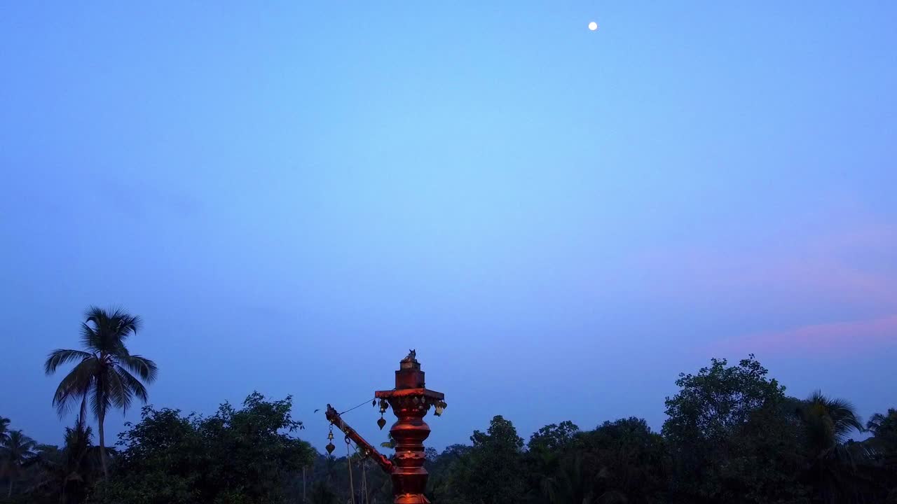 Twilight sky with moon over Indian temple lamp and palm trees. Birds fly past