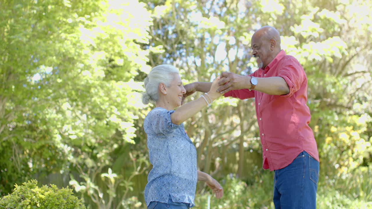 Pareja mayor birracial feliz bailando en un jardín soleado en casa, a cámara lenta