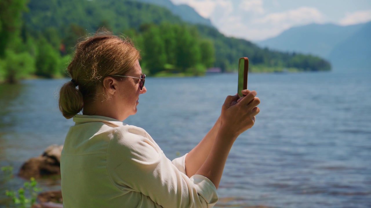 Woman stands by calm lake, phone in hand, capturing scenic view. She wears sunglasses, white shirt, hair in neat bun. Focus on peaceful moment, nature's beauty reflected in her calm posture