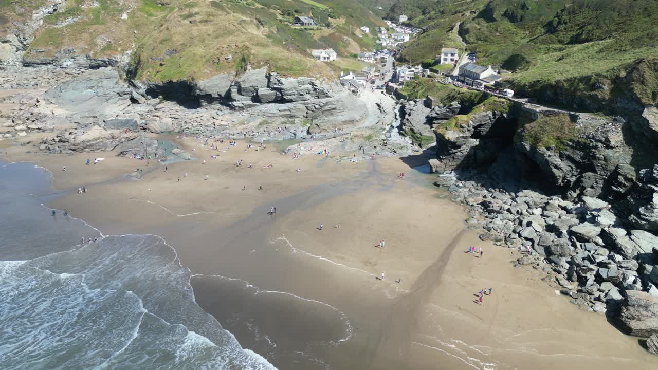 Drone footage captures Trebarwith Strand's golden sands, rugged cliffs, and azure water. Perfect sunny day with scattered beachgoers, showcasing nature's tranquil beauty