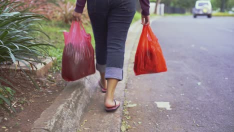 follow closeup low angle shot of a girl wearing blue jeans carrying two red plastic bags while walking at the side of the road