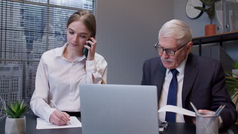 Senior-businessman-company-director-working-on-laptop-computer-in-office,-secretary-talking-on-phone