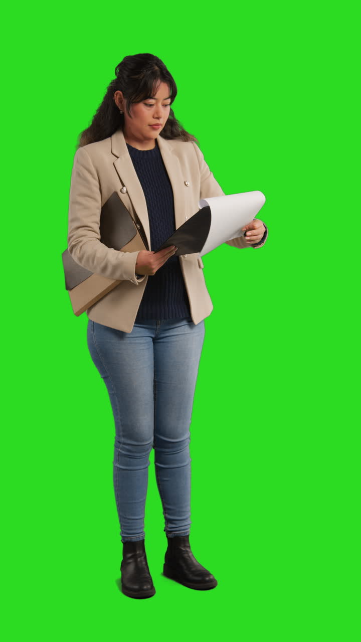 A young businesswoman stands in front of a green screen holding a document and a folder.